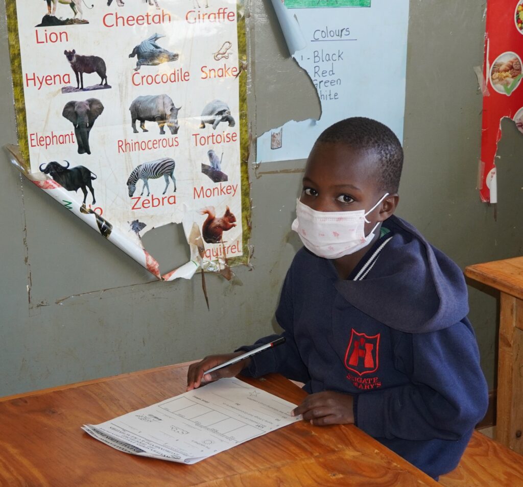 Child in Africa wearing a mask and writing at a school desk near animal posters.