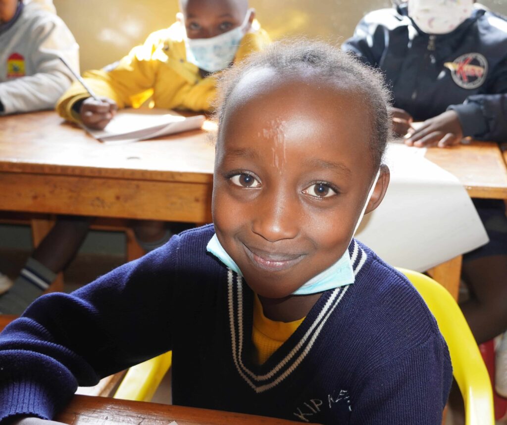 Smiling young girl in Africa sitting at a classroom desk with a mask around her neck.