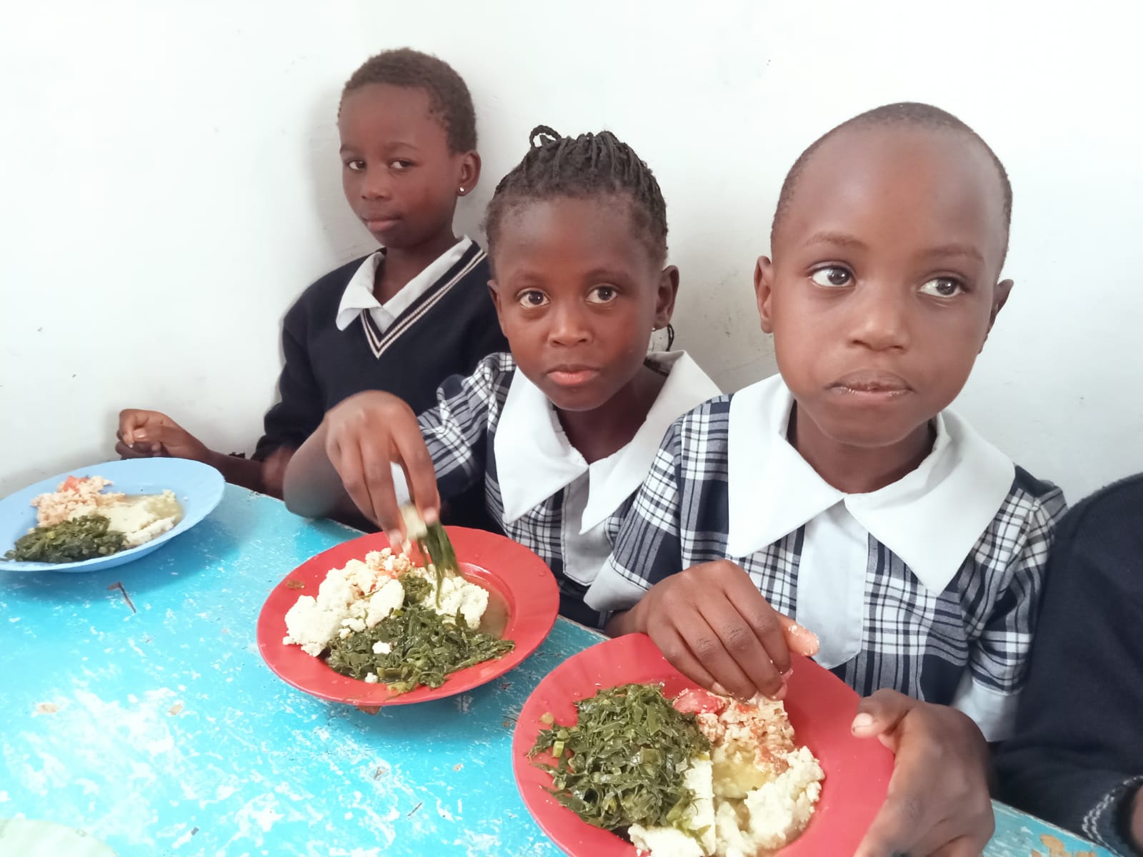 Three children in Africa eating a healthy meal of vegetables and maize at a blue table.
