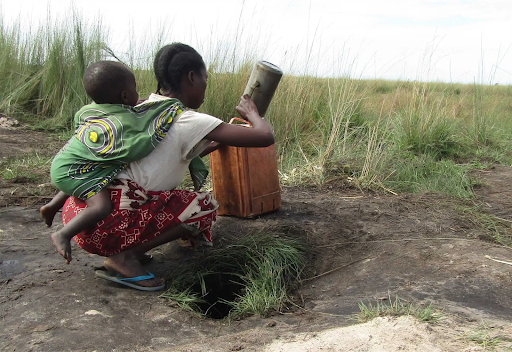 A Zambian mother with her child strapped to her back scoops water from a ground hole.